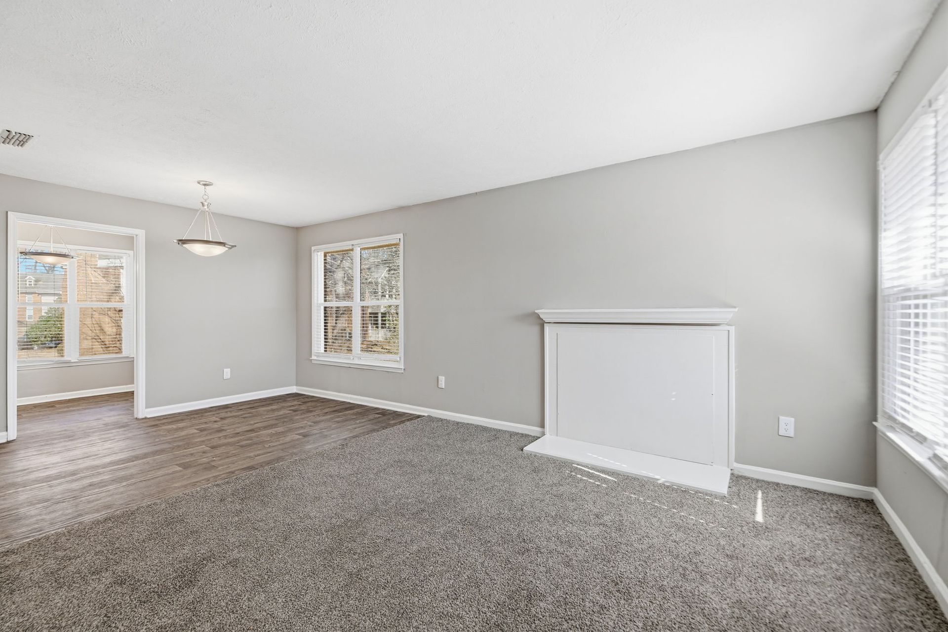A light gray living room with gray carpet, a white fireplace, and a doorway leading to a dining area with wood flooring.