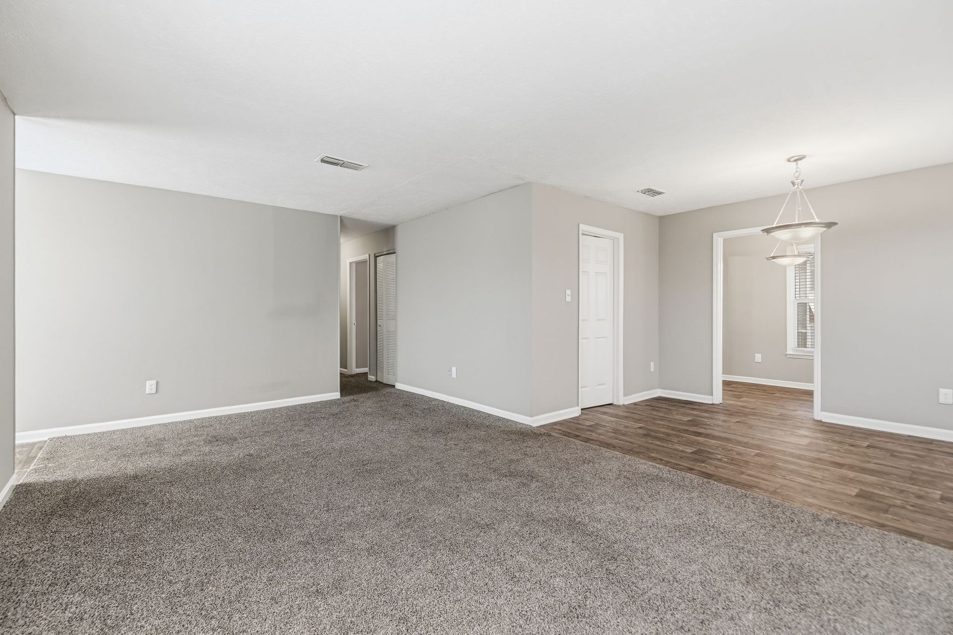 An empty room with gray carpet and walls, transitioning to a wood-floored dining area with a chandelier.