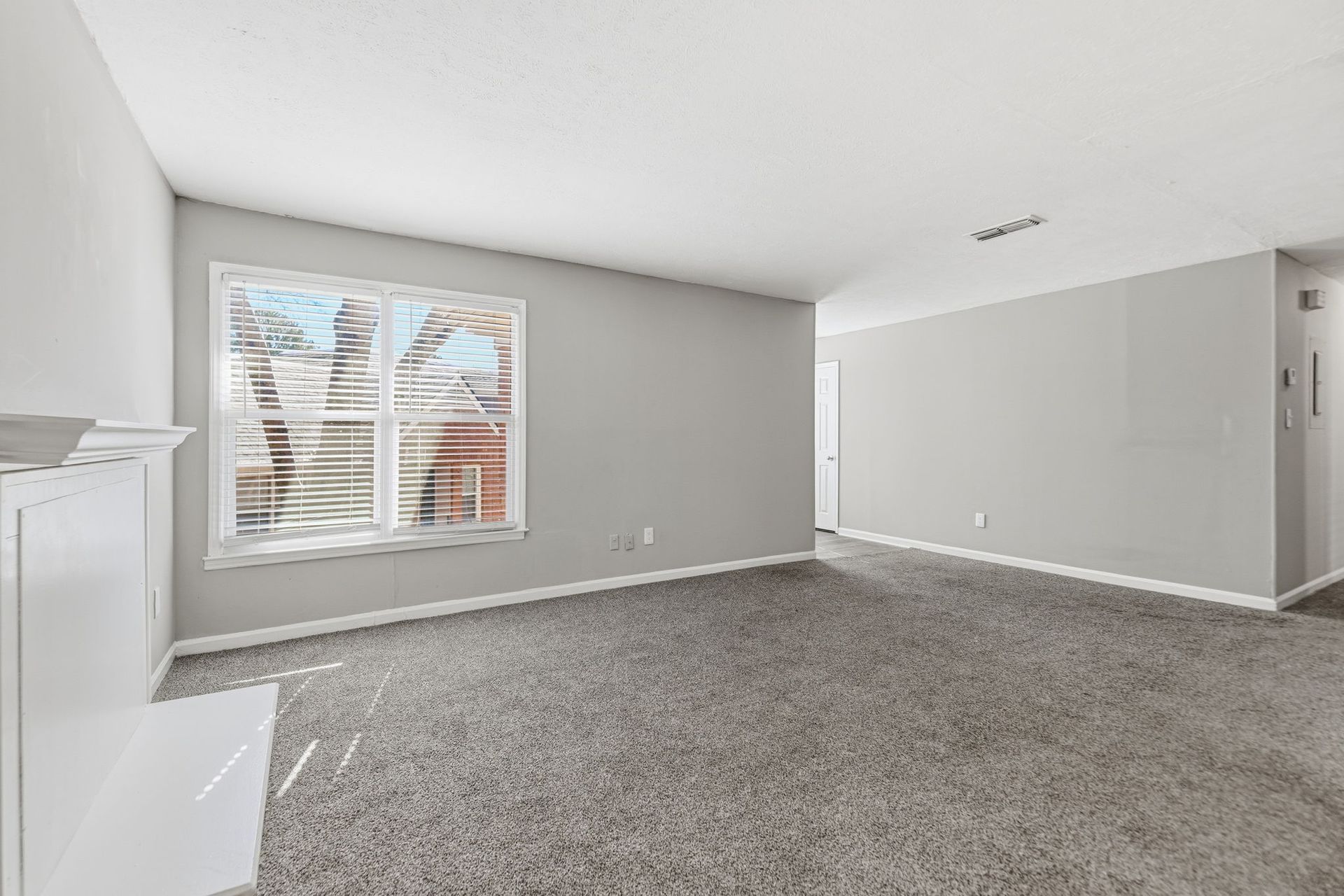 An empty living room with light gray walls, plush gray carpet, a white fireplace mantel, and a large window.