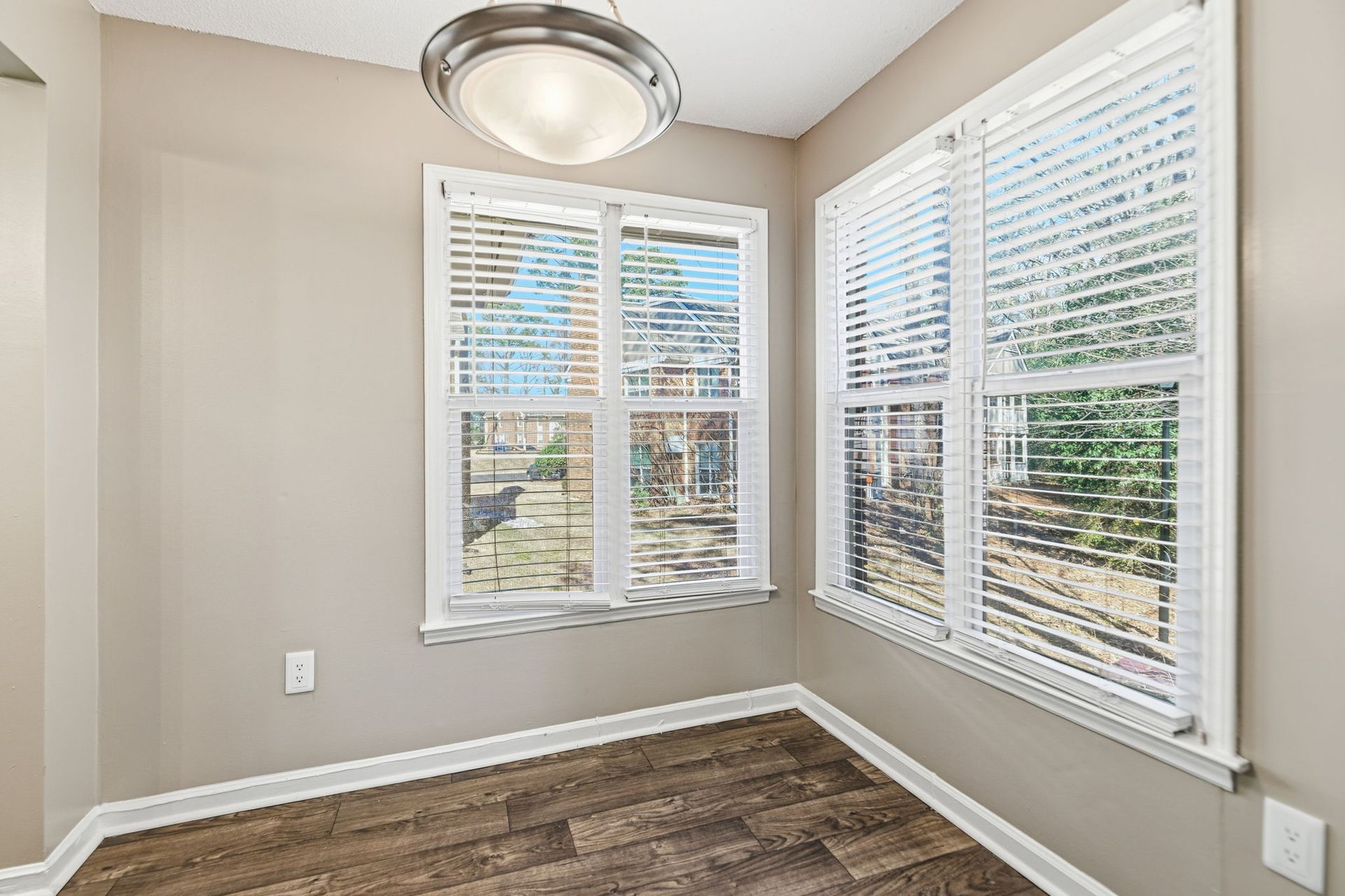 Empty room with beige walls, wood-look flooring, a modern circular light fixture, and two windows with white blinds.