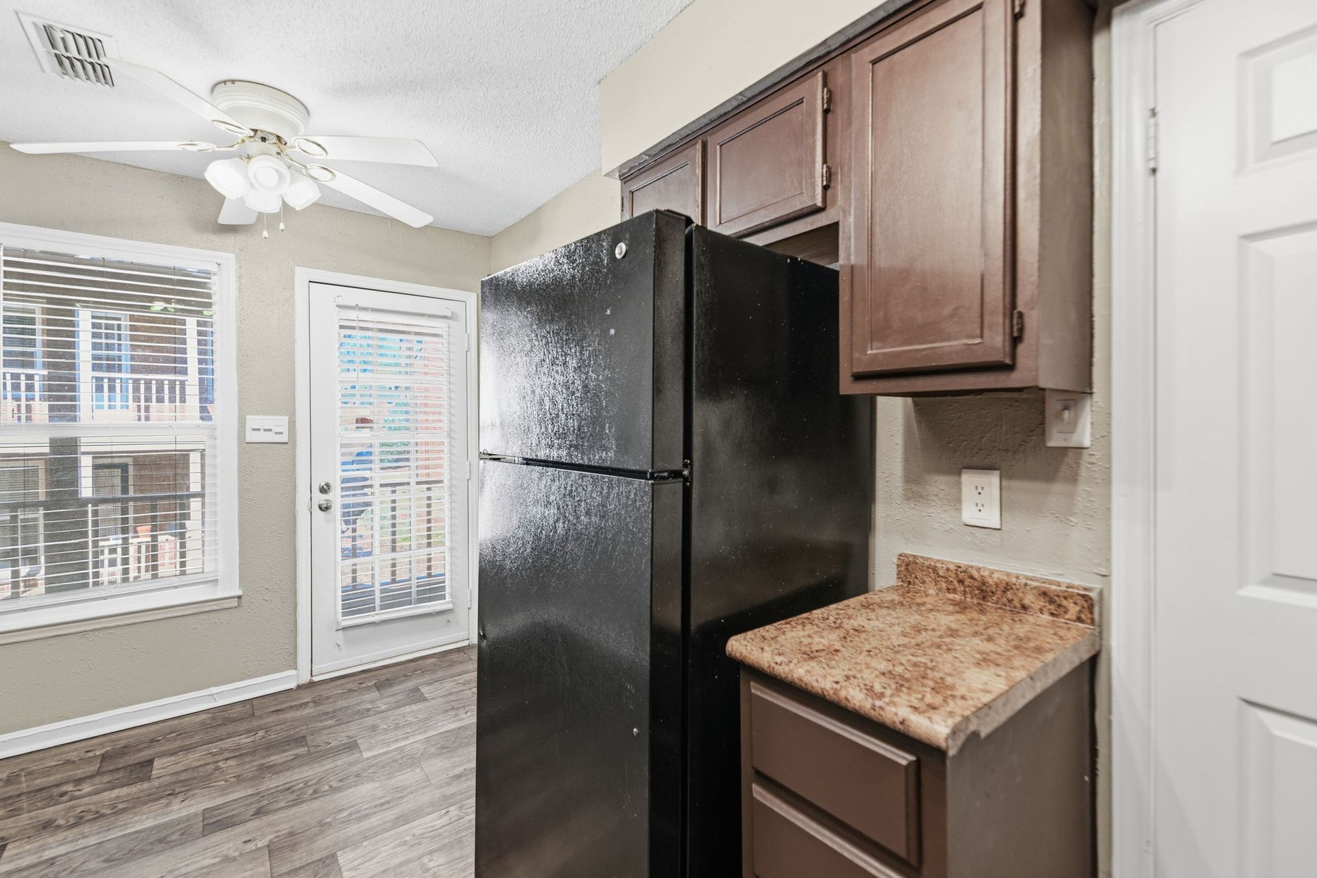 A corner of a kitchen with a tall black refrigerator, brown cabinets, patterned countertops, and a glass-paneled door.