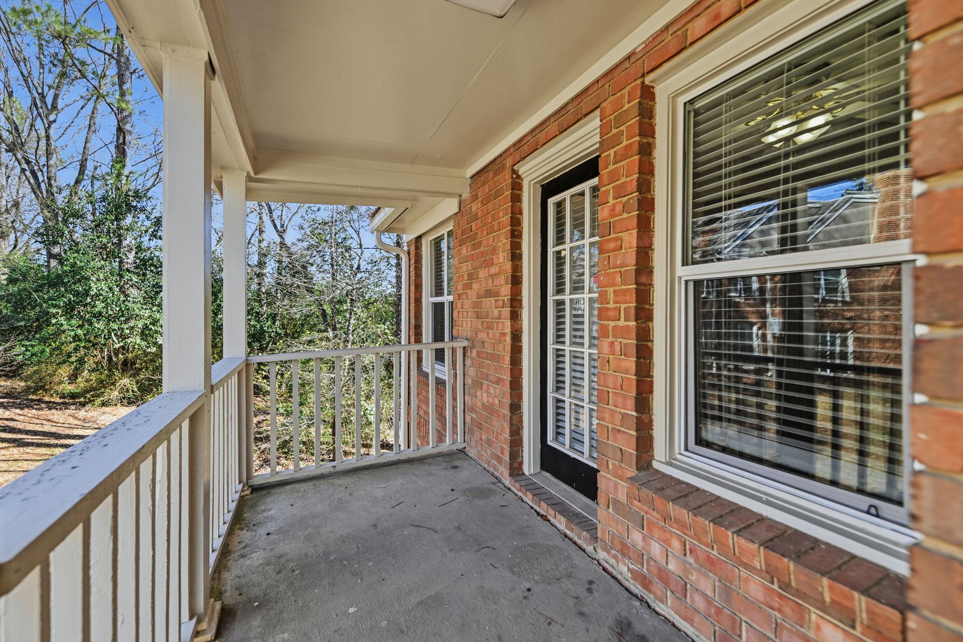 A covered brick porch with a white railing and a glass door, looking out over a wooded area on a sunny day.