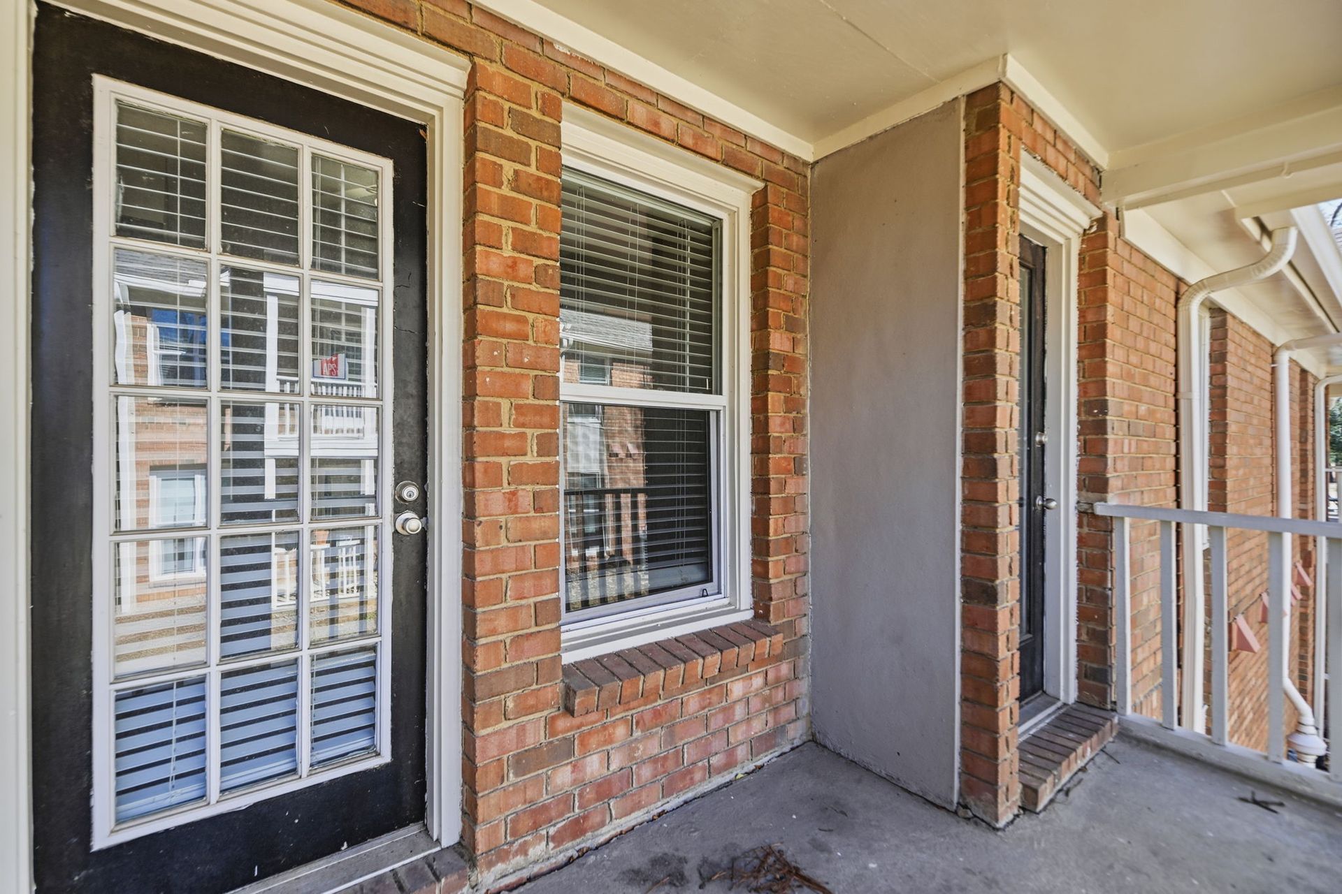A brick apartment patio featuring a black door with glass panels, a window with blinds, and a white wooden railing.