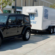 A black Jeep Wrangler parked on a concrete surface, towing a white refrigerated trailer labeled