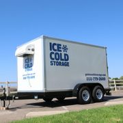 A white portable refrigerator trailer parked on pavement against a clear blue sky, displaying 