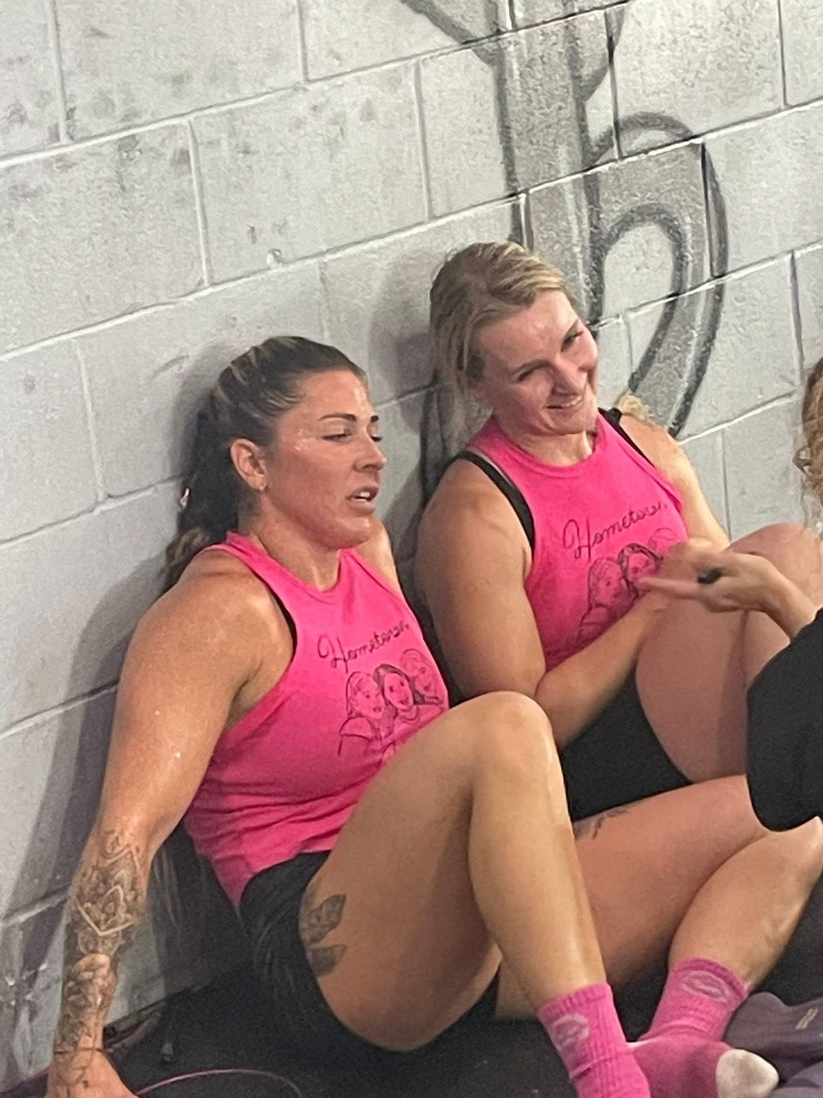 Two people in matching pink tank tops sit on the floor against a cinderblock wall, resting after a workout.