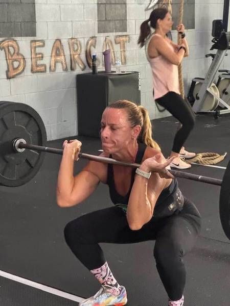 A person in a gym in Bunnell, FL performs a barbell front squat, while another person in the background climbs a hanging rope.