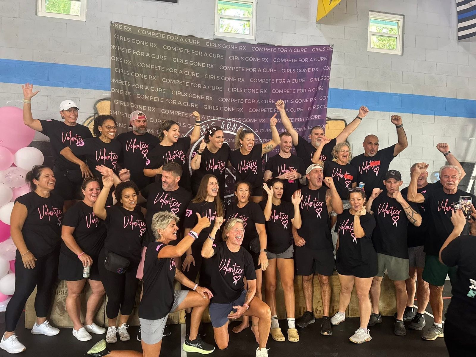 A large group of people in matching black shirts pose together indoors in front of a purple banner with white text.