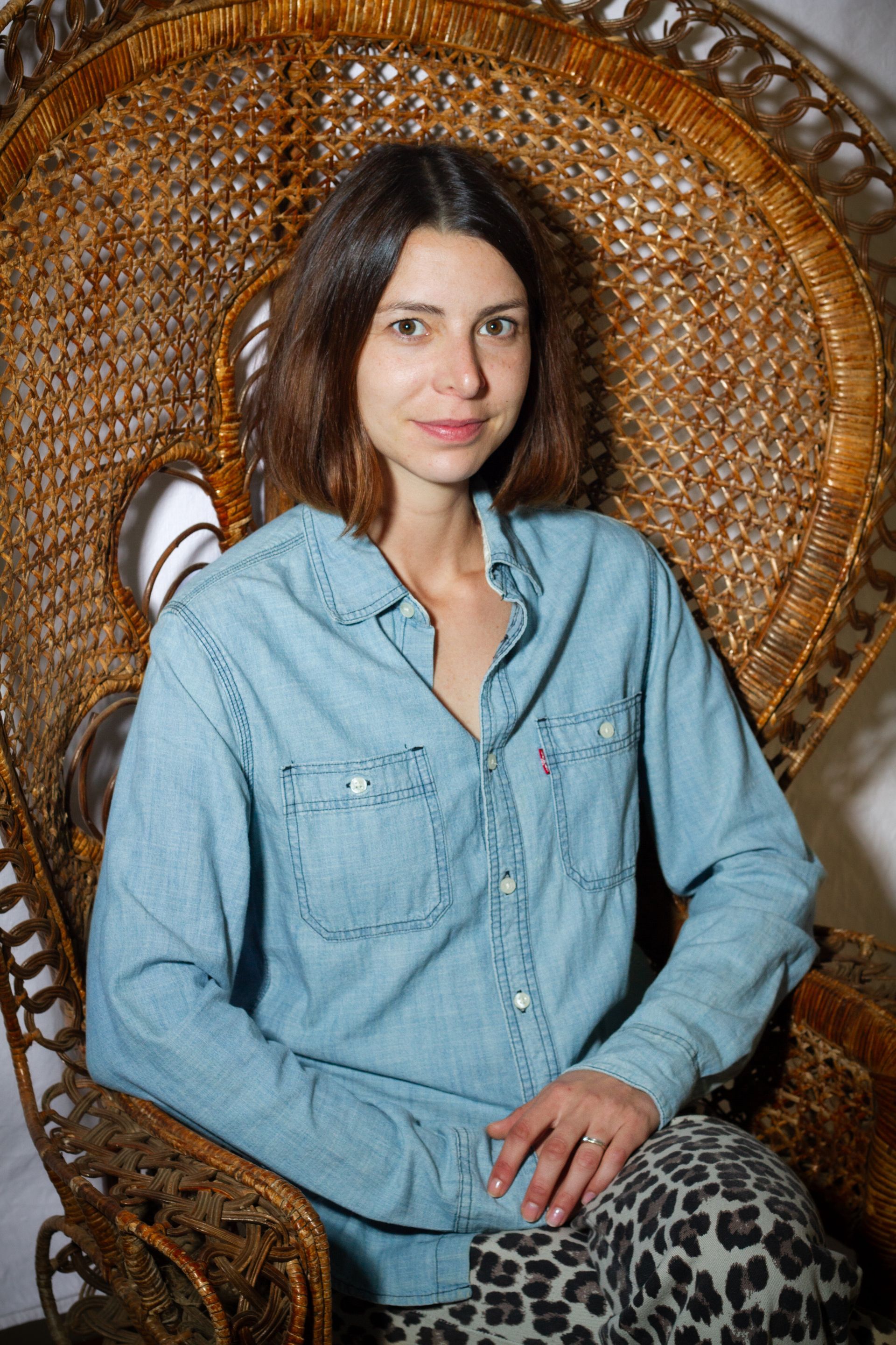 Une femme en chemise en jean est assise dans un fauteuil en osier orné.