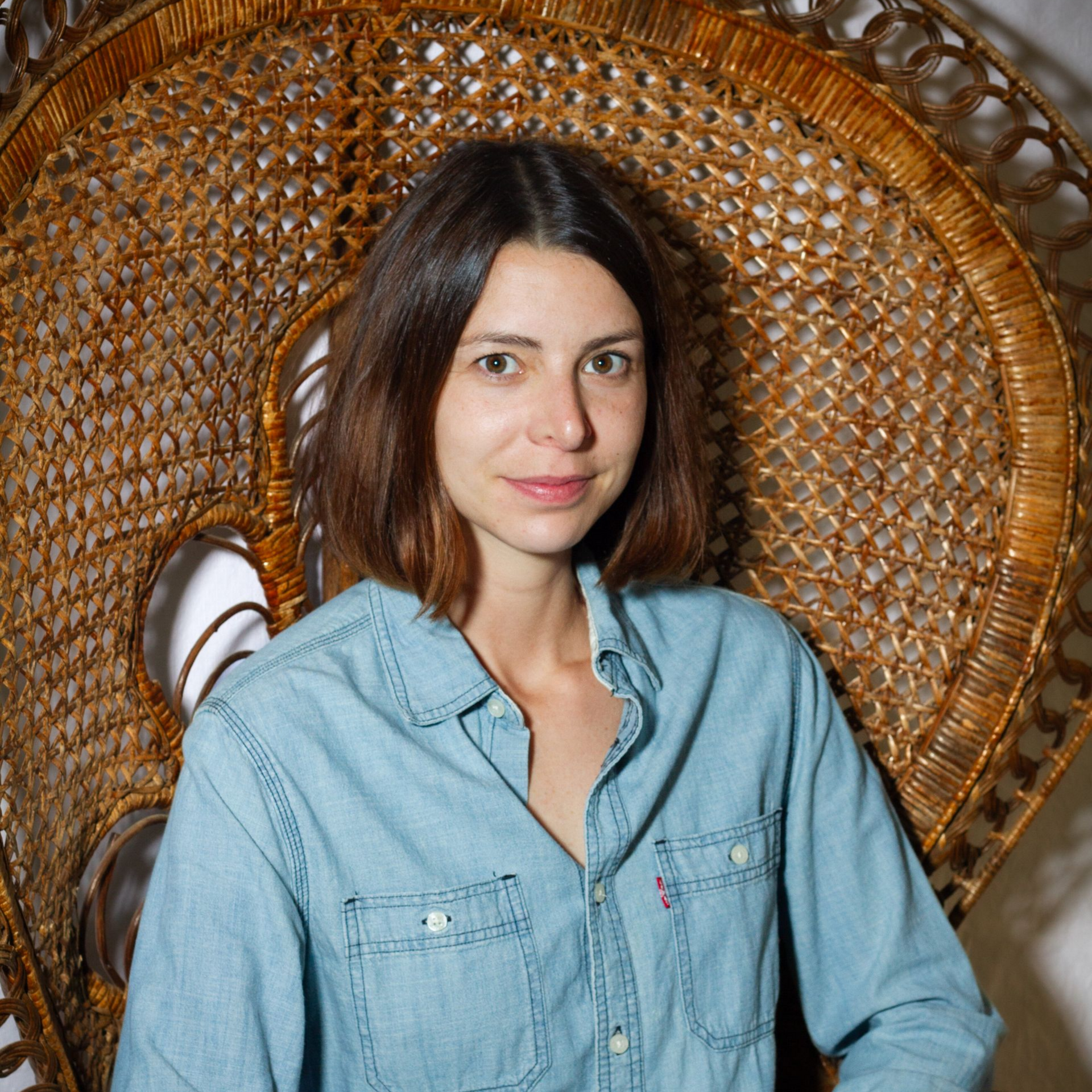 Une femme en chemise en jean est assise dans un fauteuil en osier orné.