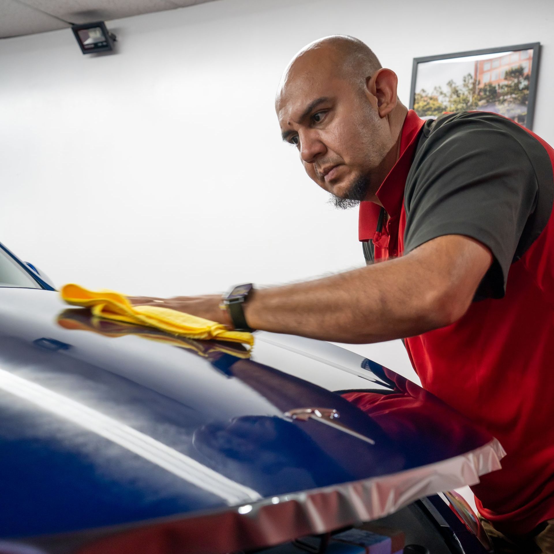 A man in a red shirt is cleaning a blue car with a yellow cloth