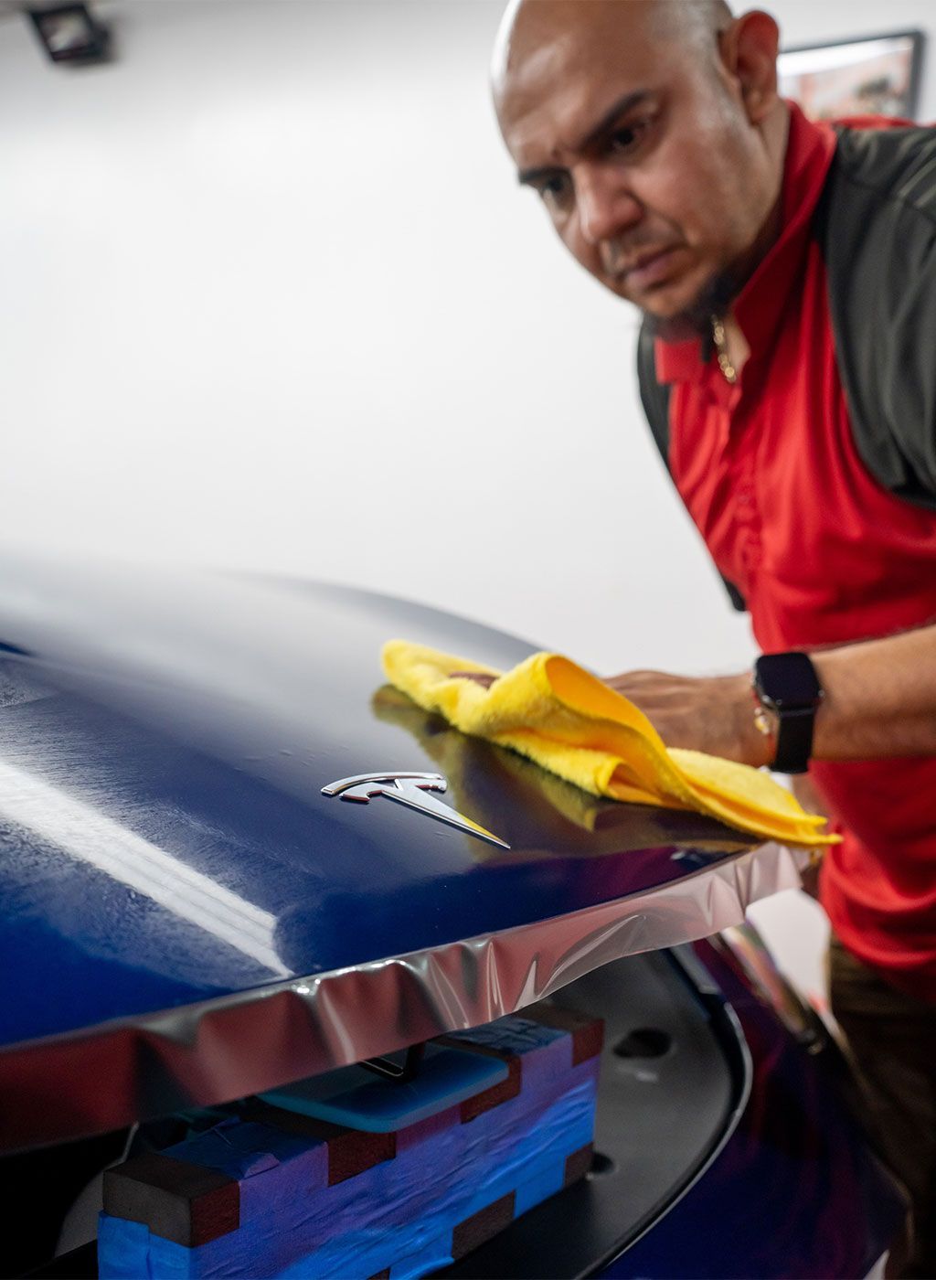 A man in a red shirt is cleaning a blue car with a yellow cloth