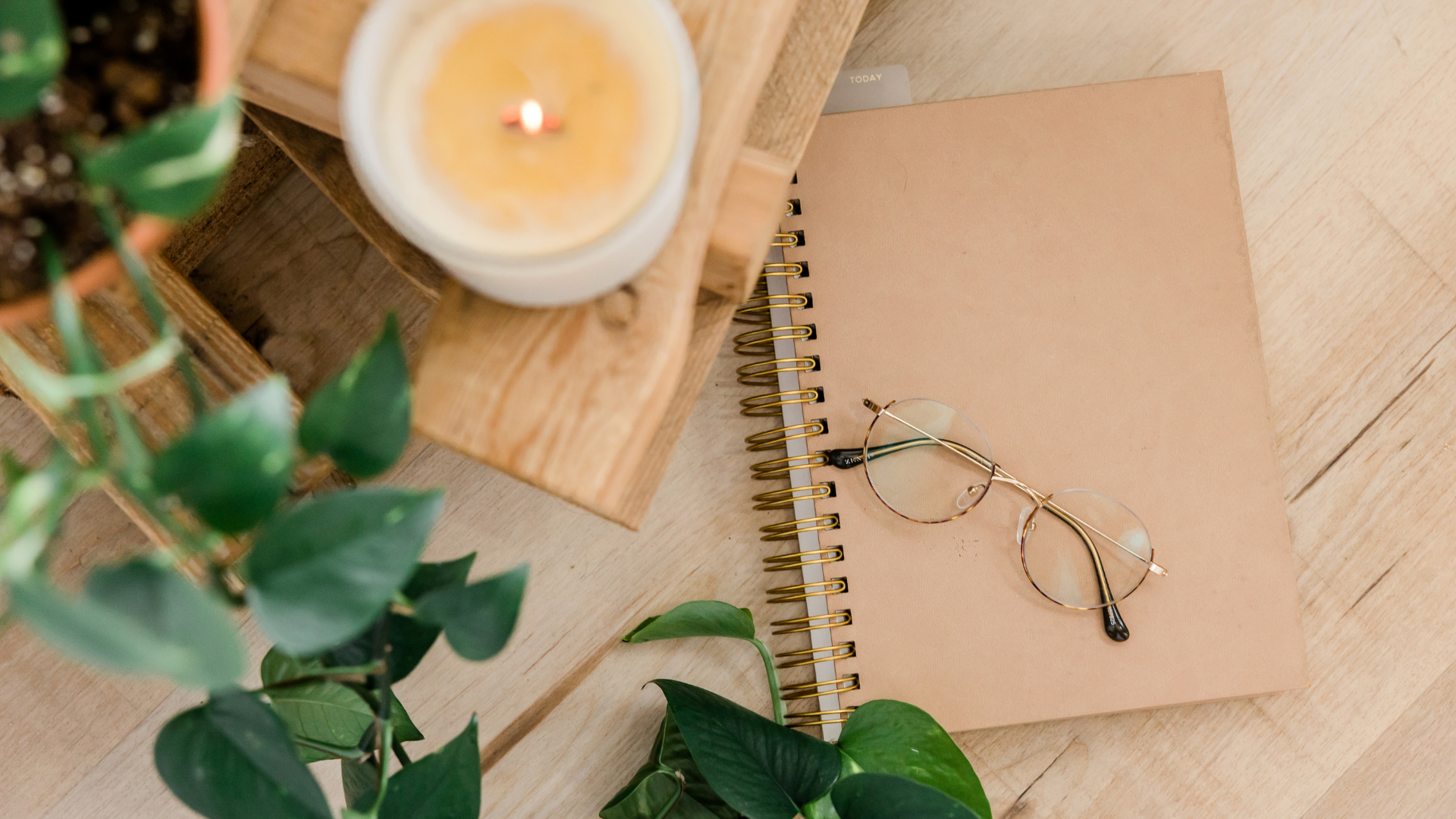 A candle , notebook , glasses and a plant are on a wooden table.