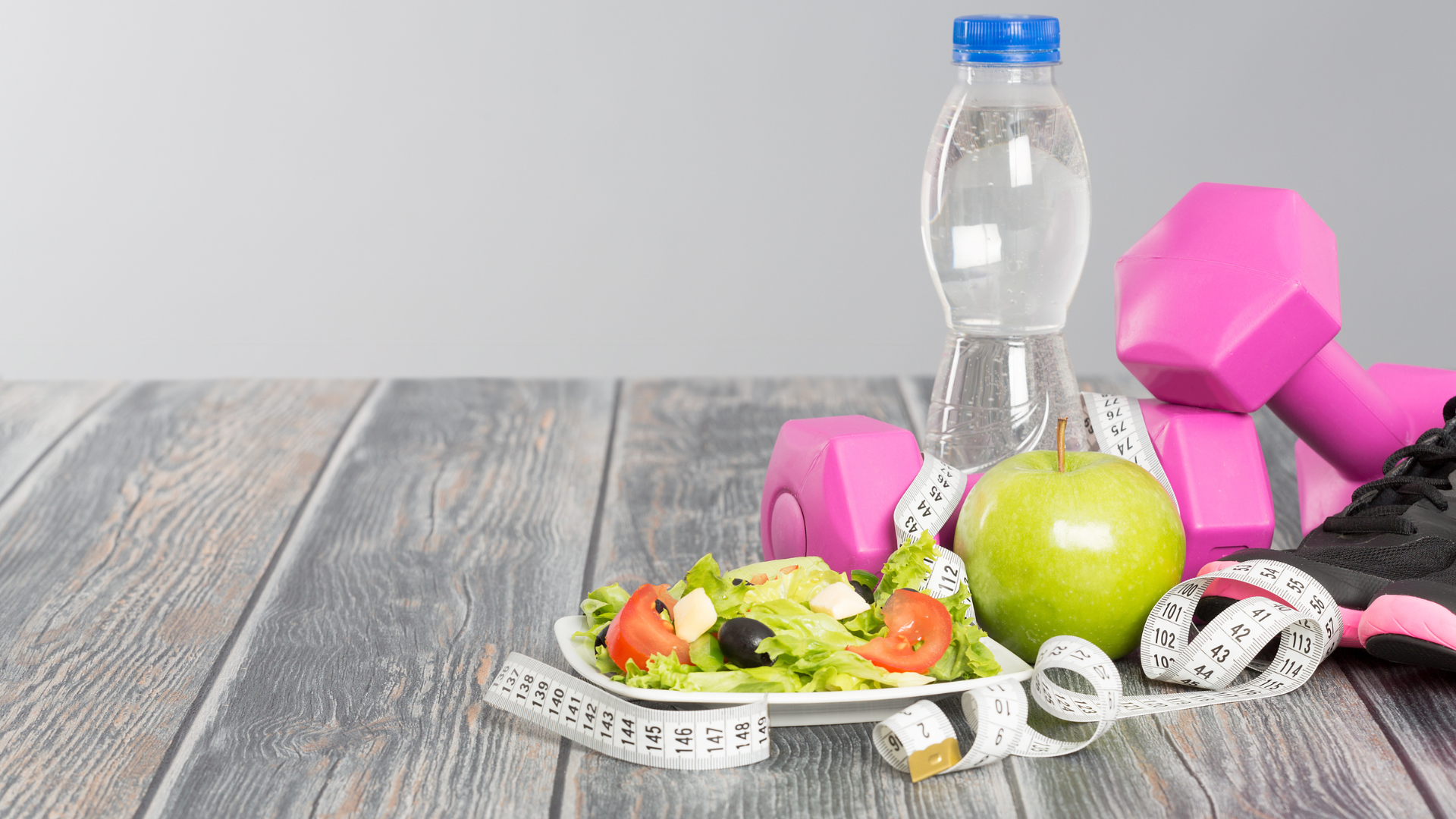 A plate of food , dumbbells , a bottle of water and a measuring tape on a wooden table.