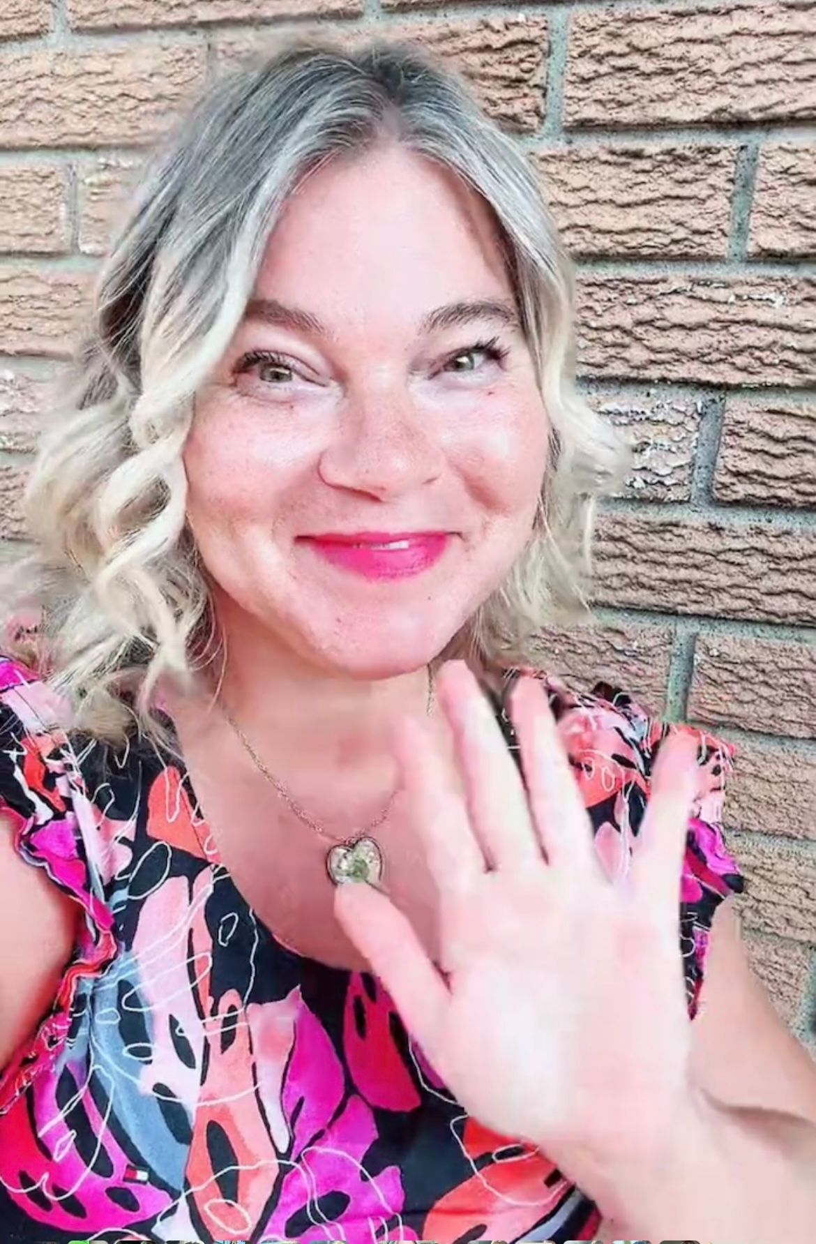 A woman is waving her hand in front of a brick wall.