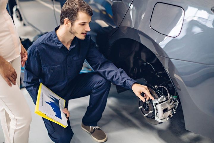 Mechanic kneeling beside a gray car, inspecting exposed front wheel area with a clipboard nearby