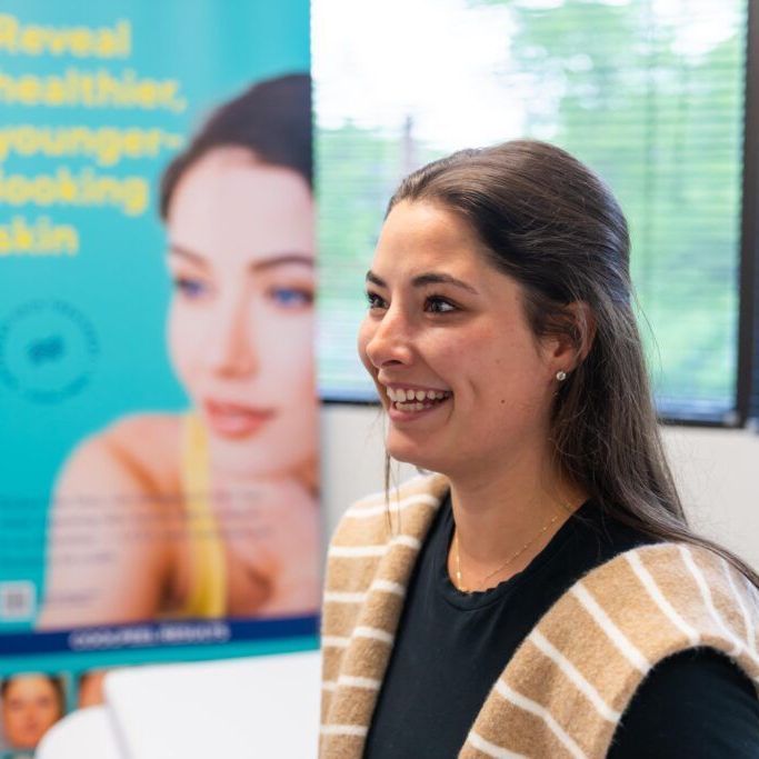 A woman is smiling in front of a sign that says reveal healthier younger looking skin