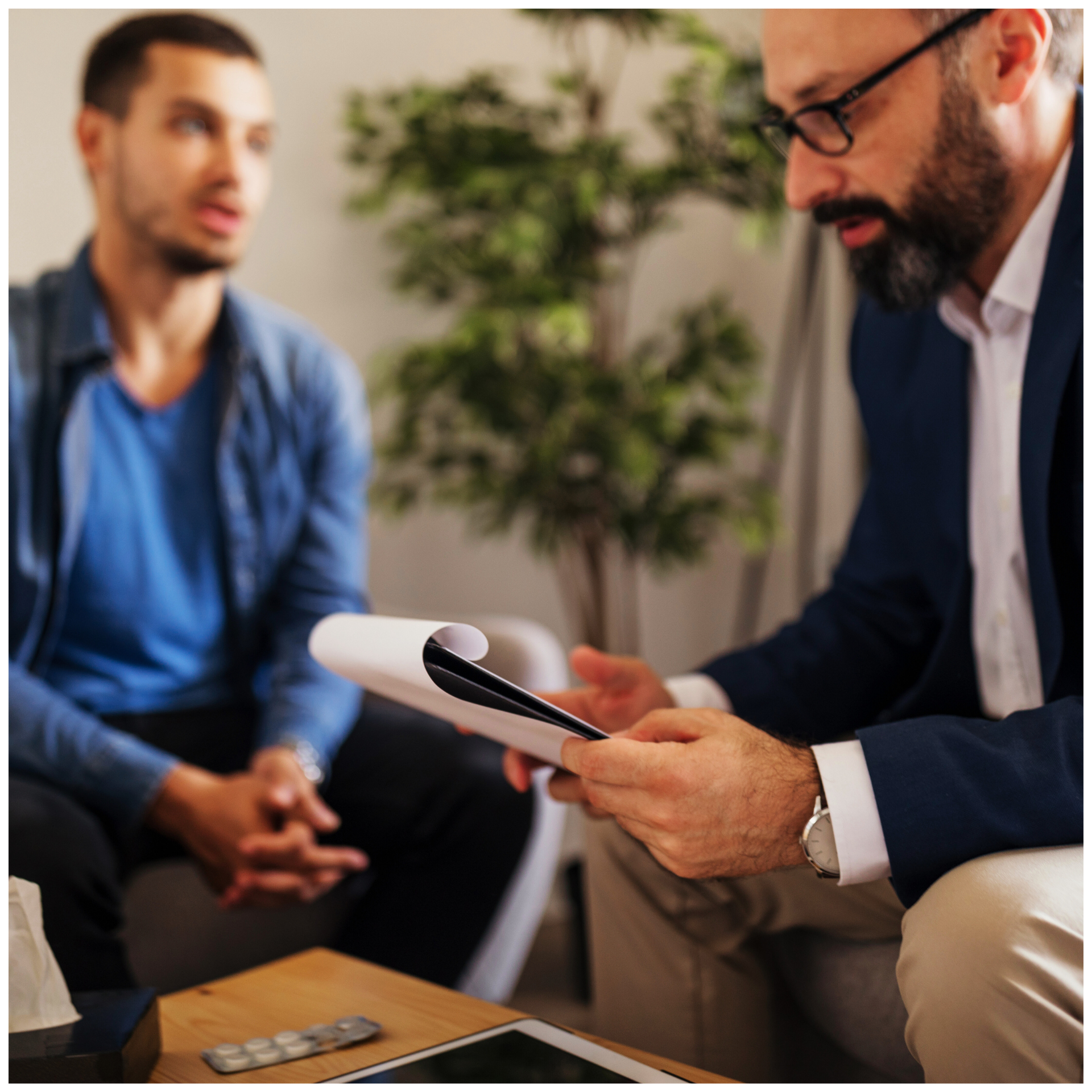 A man is sitting on a couch talking to another man while holding a piece of paper.