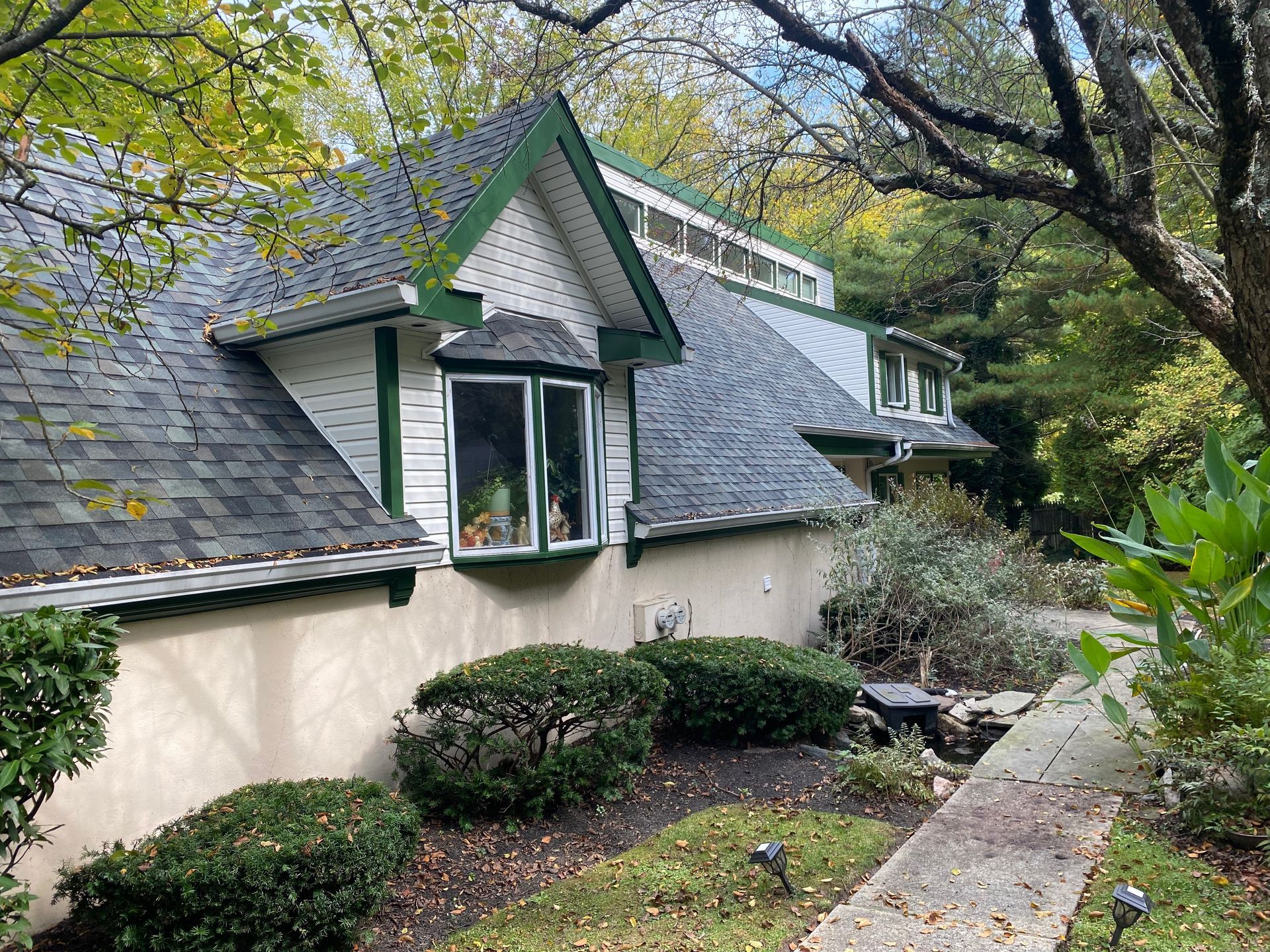 A white house with a green trim and a black roof is surrounded by trees and bushes.