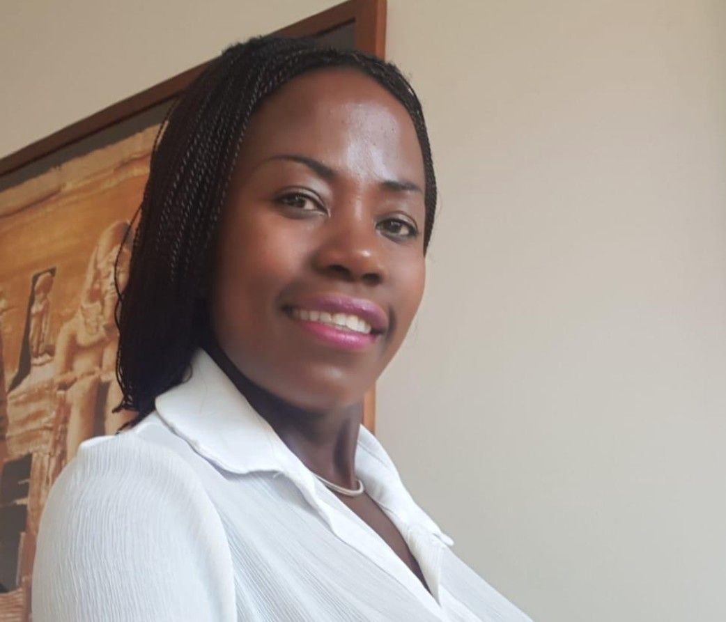 Smilende sort kvinde med flettet hår iført en hvid skjorte med krave i et rum med et indrammet kunstværk.