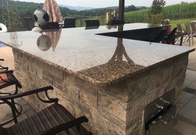 Outdoor kitchen island with a stone base and polished granite countertop, set on a patio with bar stools and a grill.