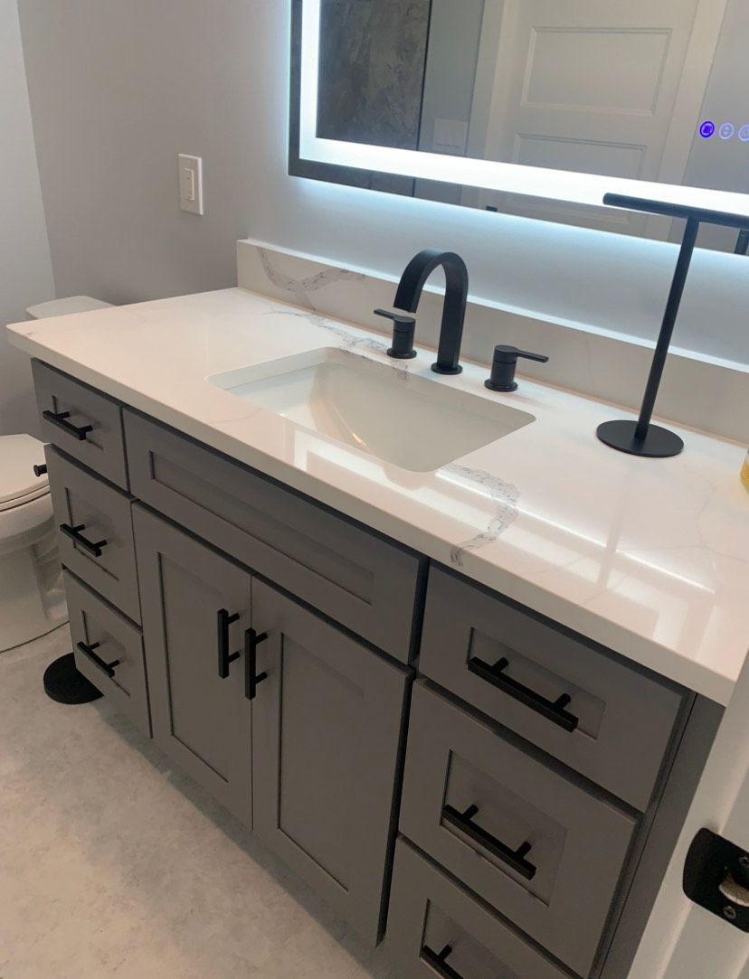 A modern bathroom vanity with a gray cabinet, white stone countertop, black faucet, and a large backlit wall mirror.