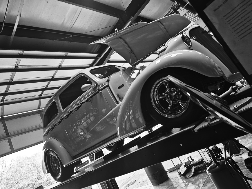 Classic car on a lift in a garage, viewed from below with its hood open. | Cruz N Carz