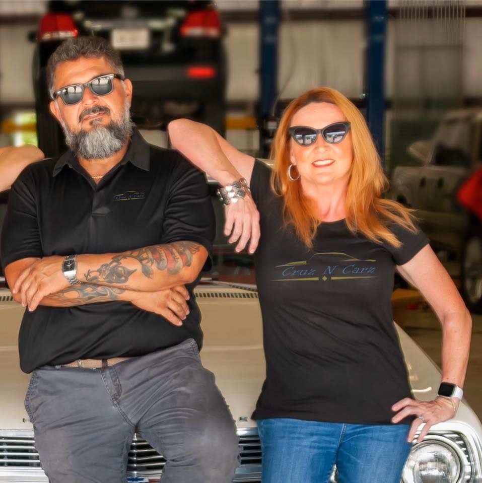 Two people in sunglasses leaning on a car in a garage, smiling at the camera. | Cruz-N-Carz