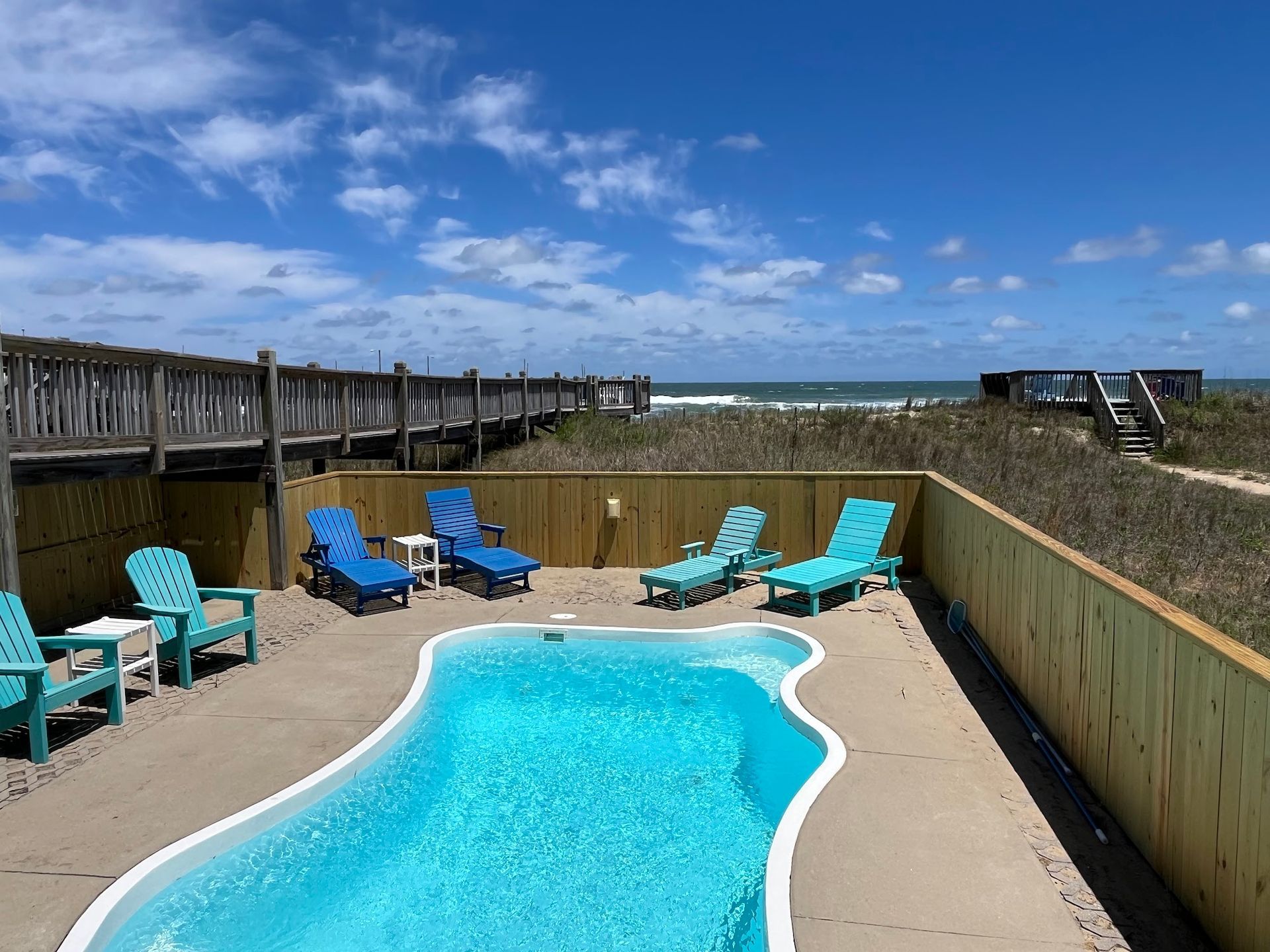 Poolside view of a beach with lounge chairs, blue water, and a boardwalk on a sunny day.