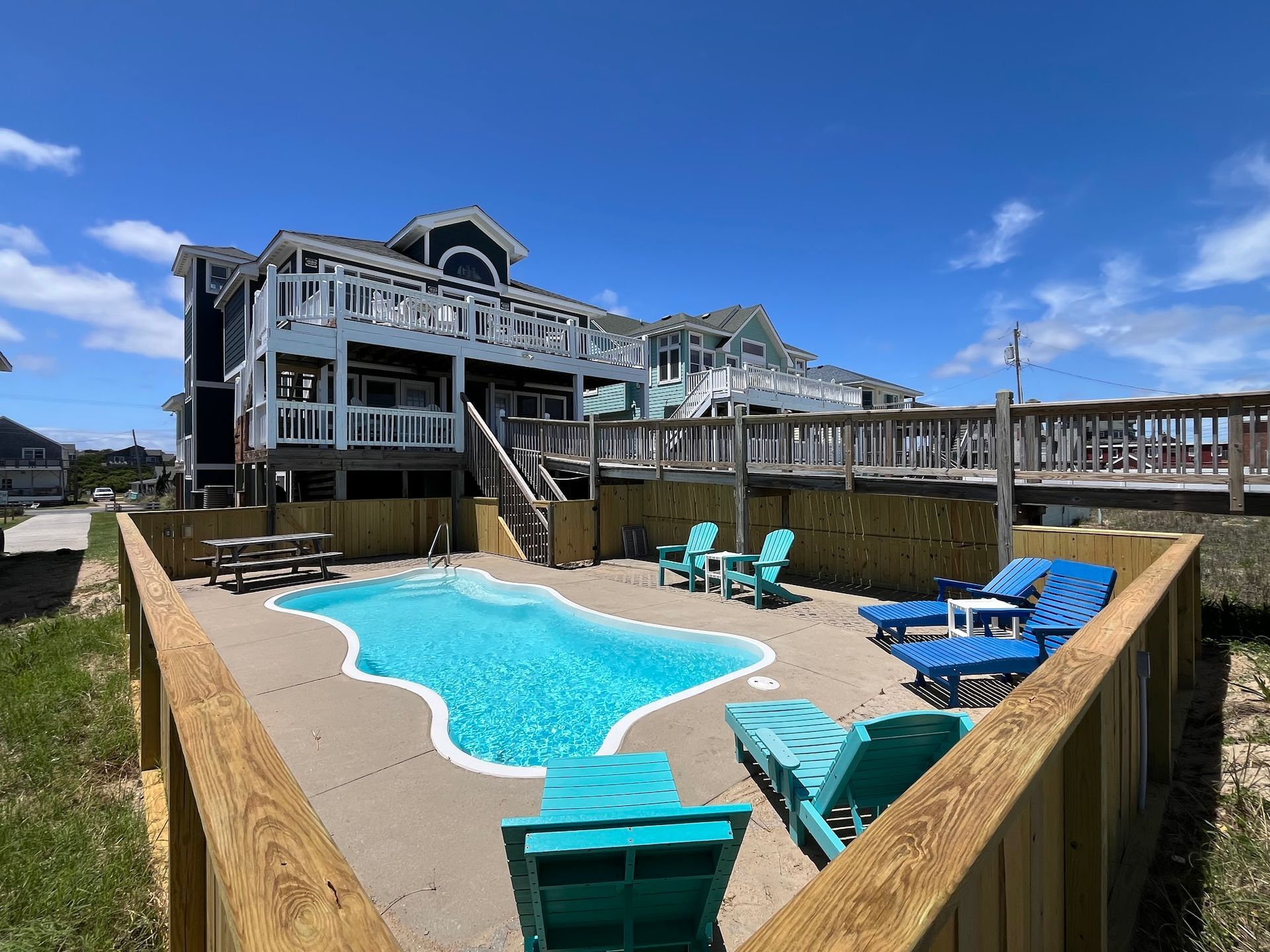 Beach house with a pool, lounge chairs, and wooden deck under a blue sky.