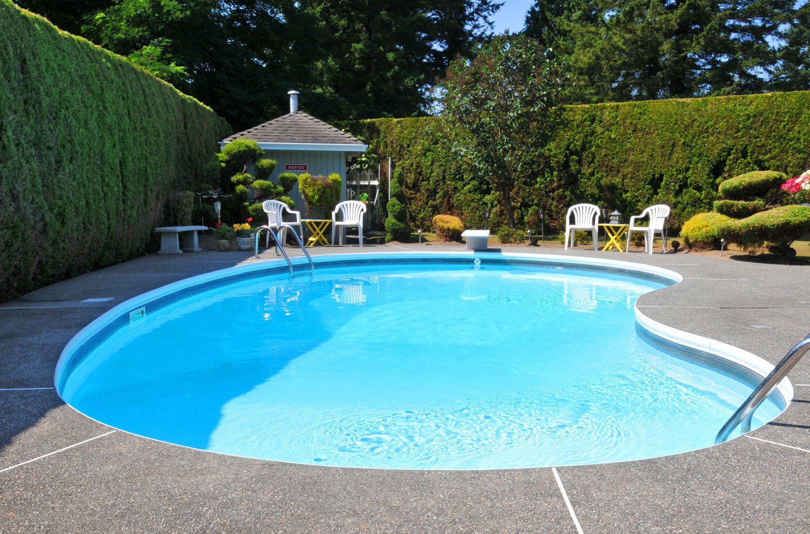 A kidney-shaped swimming pool with blue water, surrounded by concrete and lawn. White chairs sit nearby.