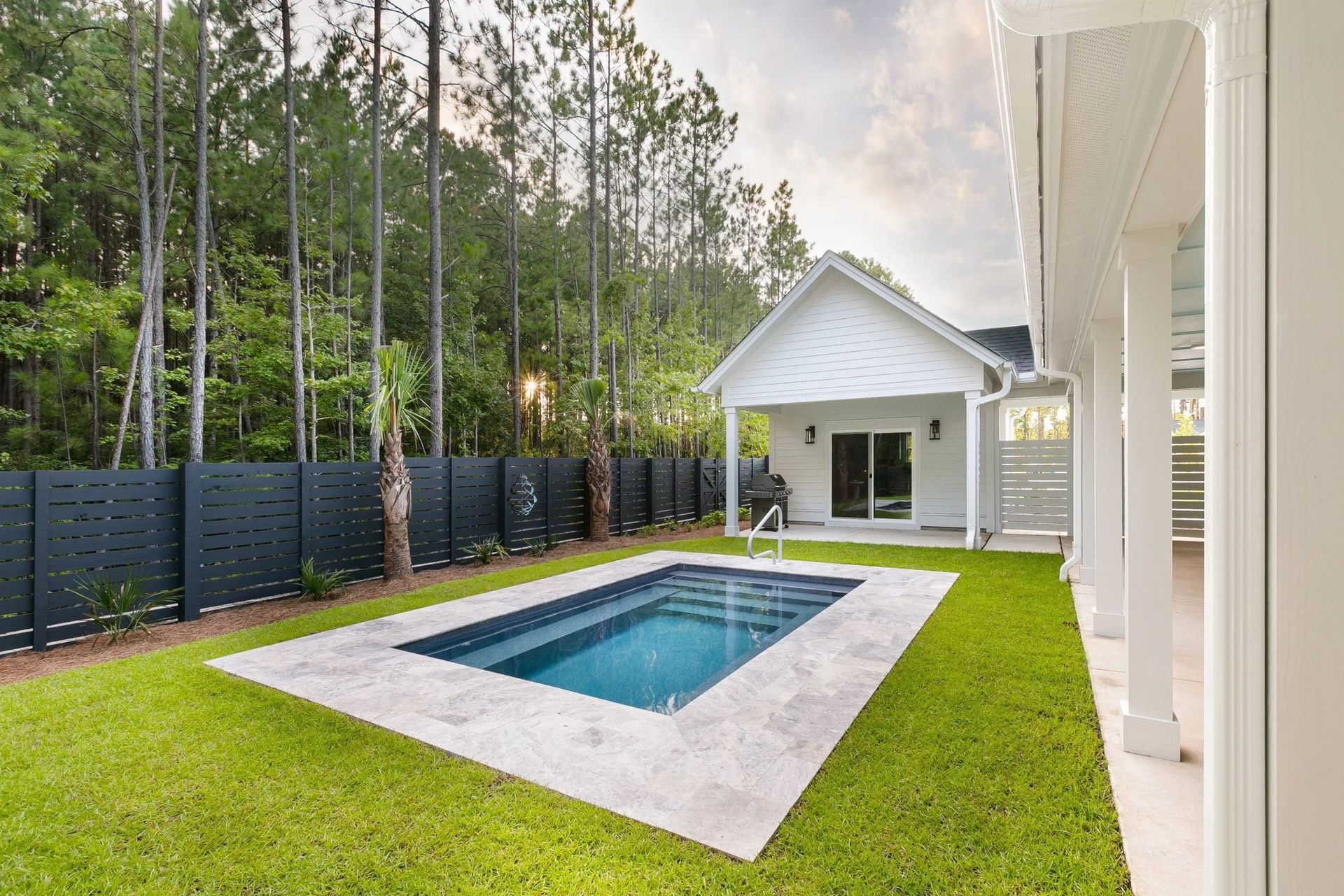 Backyard with small pool, green lawn, white house, and dark gray fence.