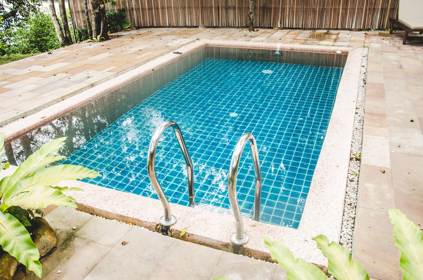 Small rectangular outdoor swimming pool with blue tiles, stainless steel ladder, and wood decking.