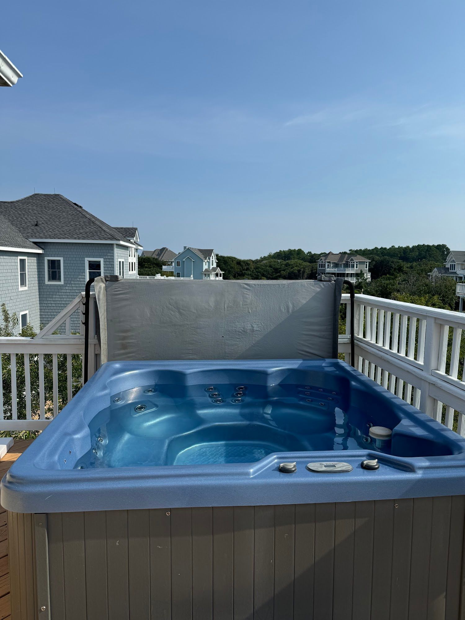 Hot tub on a wooden deck with a white railing, overlooking houses and a blue sky.