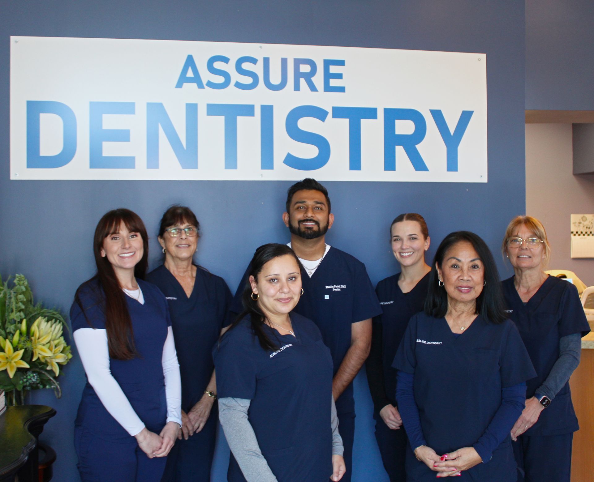 A dental team in navy scrubs poses in front of an Assure Dentistry office sign.