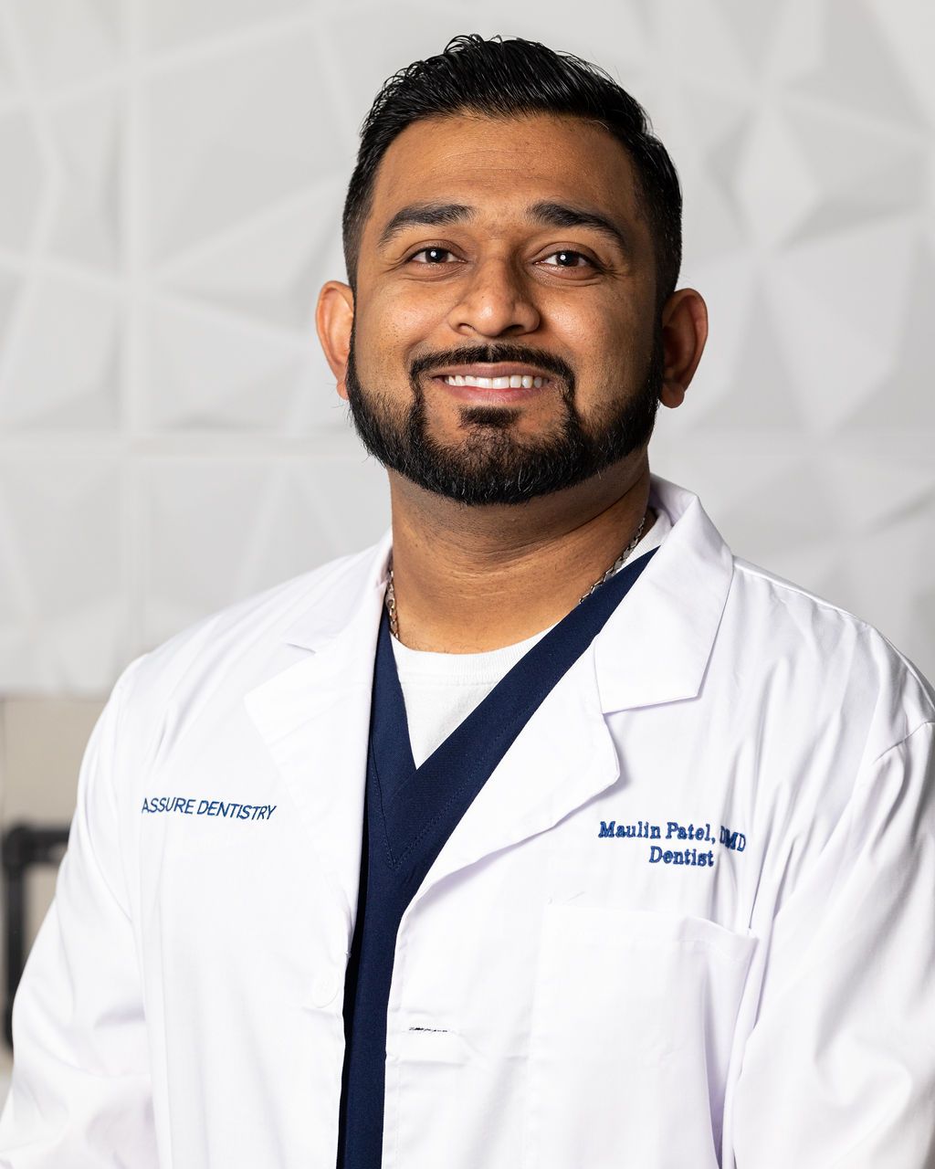 A smiling professional wearing a white lab coat and dark scrubs against a textured white background.