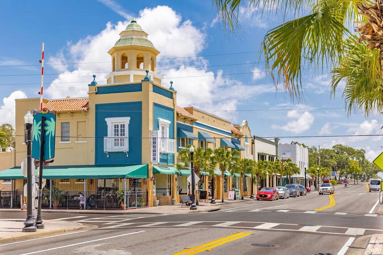Street view of buildings with colorful facades, awnings, and palm trees, under a blue sky.