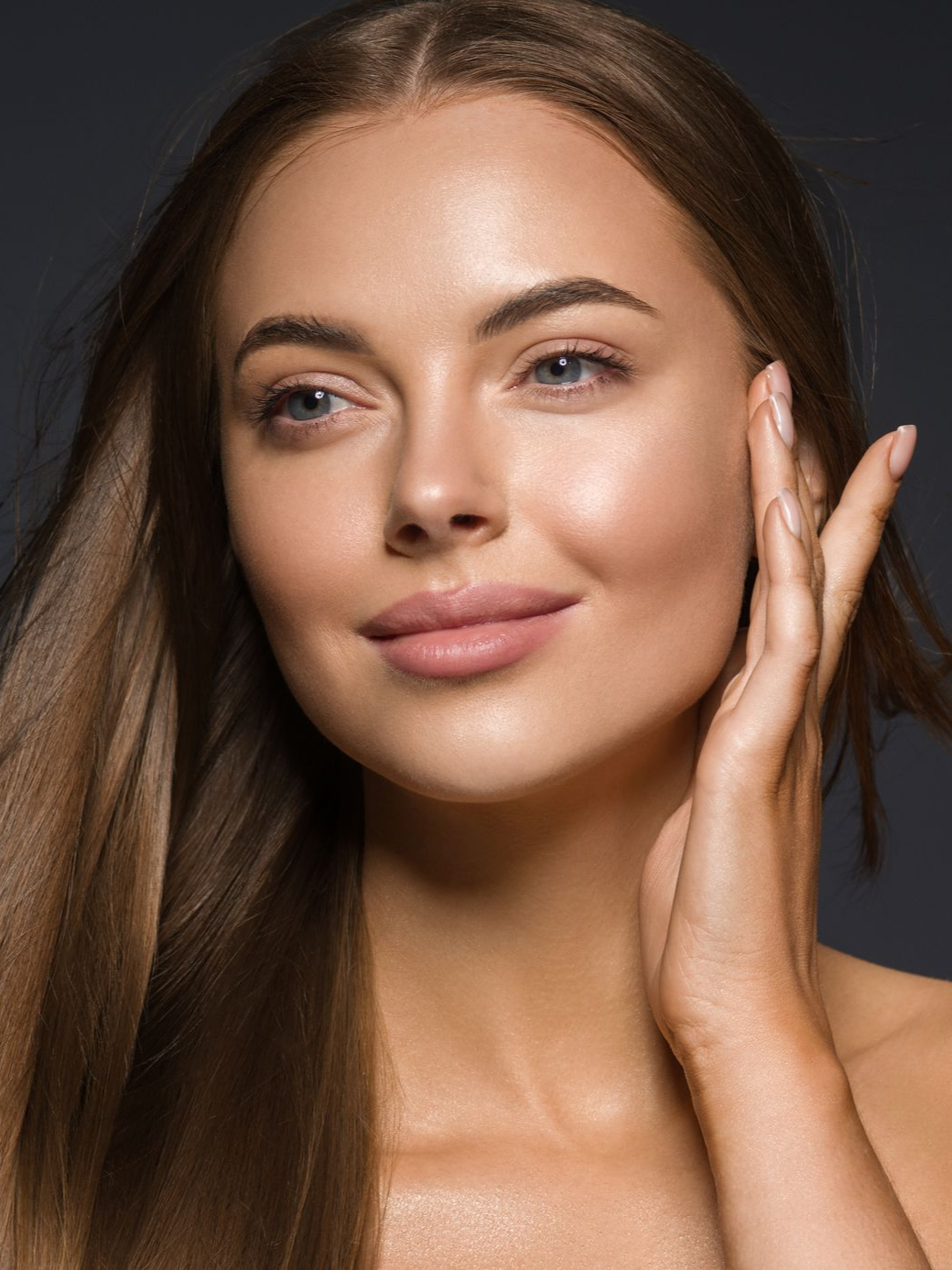 Woman with long brown hair, touching her cheek, smiling, with a soft focus and natural makeup.