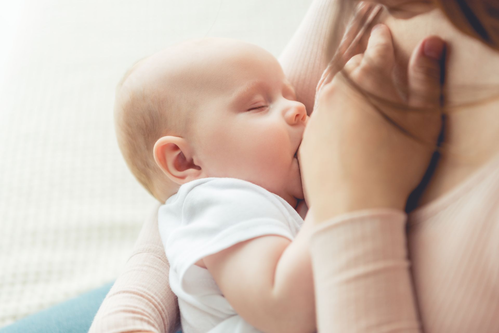 Baby nursing from a person. Baby is wearing a white onesie, eyes closed. Person's arm is wrapped around baby.