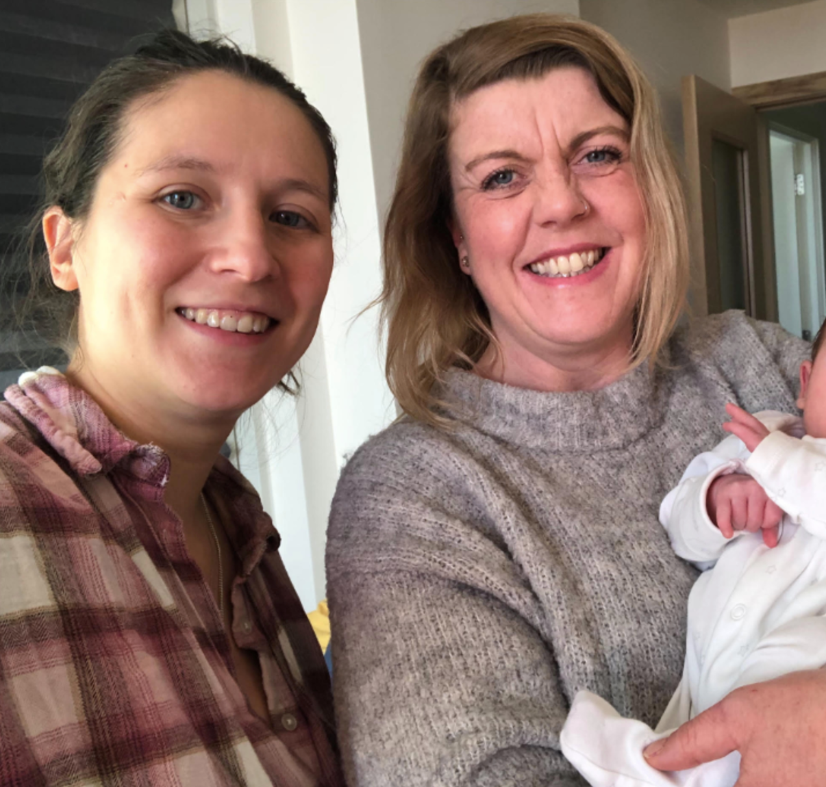 Two women smiling, one holding a baby. Indoors, natural light. One woman wears a flannel shirt, the other a sweater.