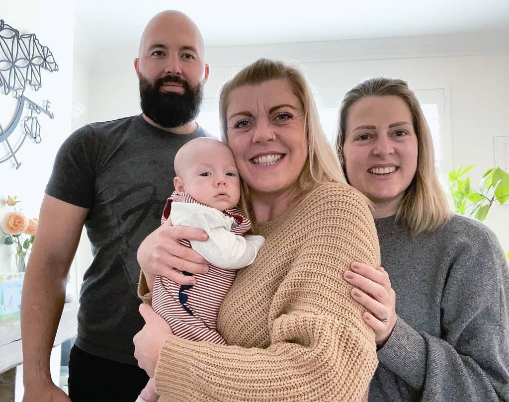 Family of four: man, woman holding baby, and another woman smiling indoors.