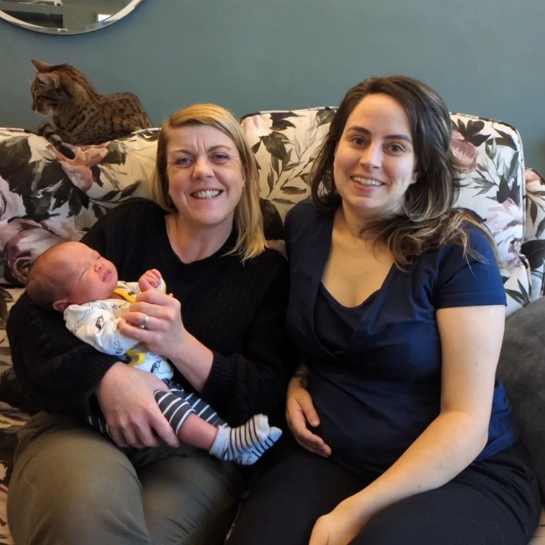Two women and a baby on a floral couch; one woman holds the infant. A cat sits behind them.