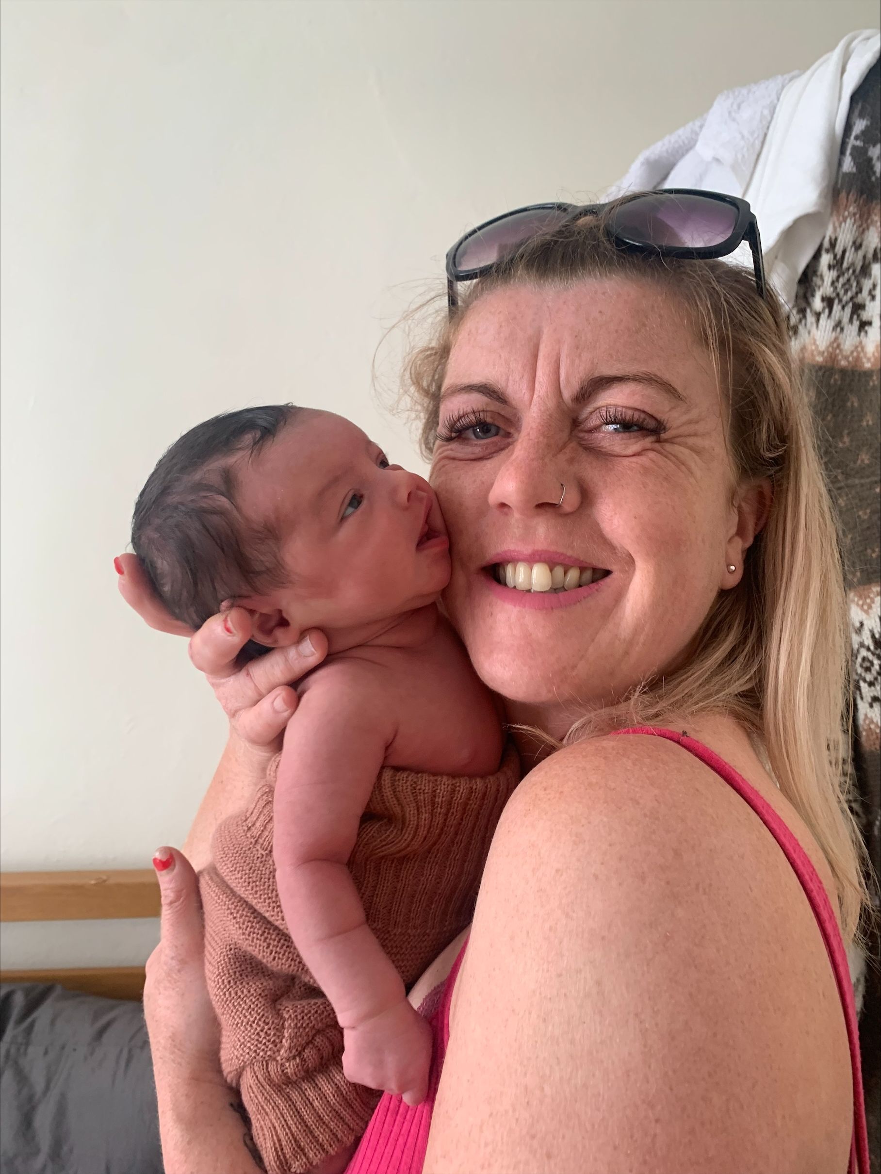 Woman holding a newborn baby; both smiling. The baby is wrapped in a brown blanket. Woman is wearing sunglasses.
