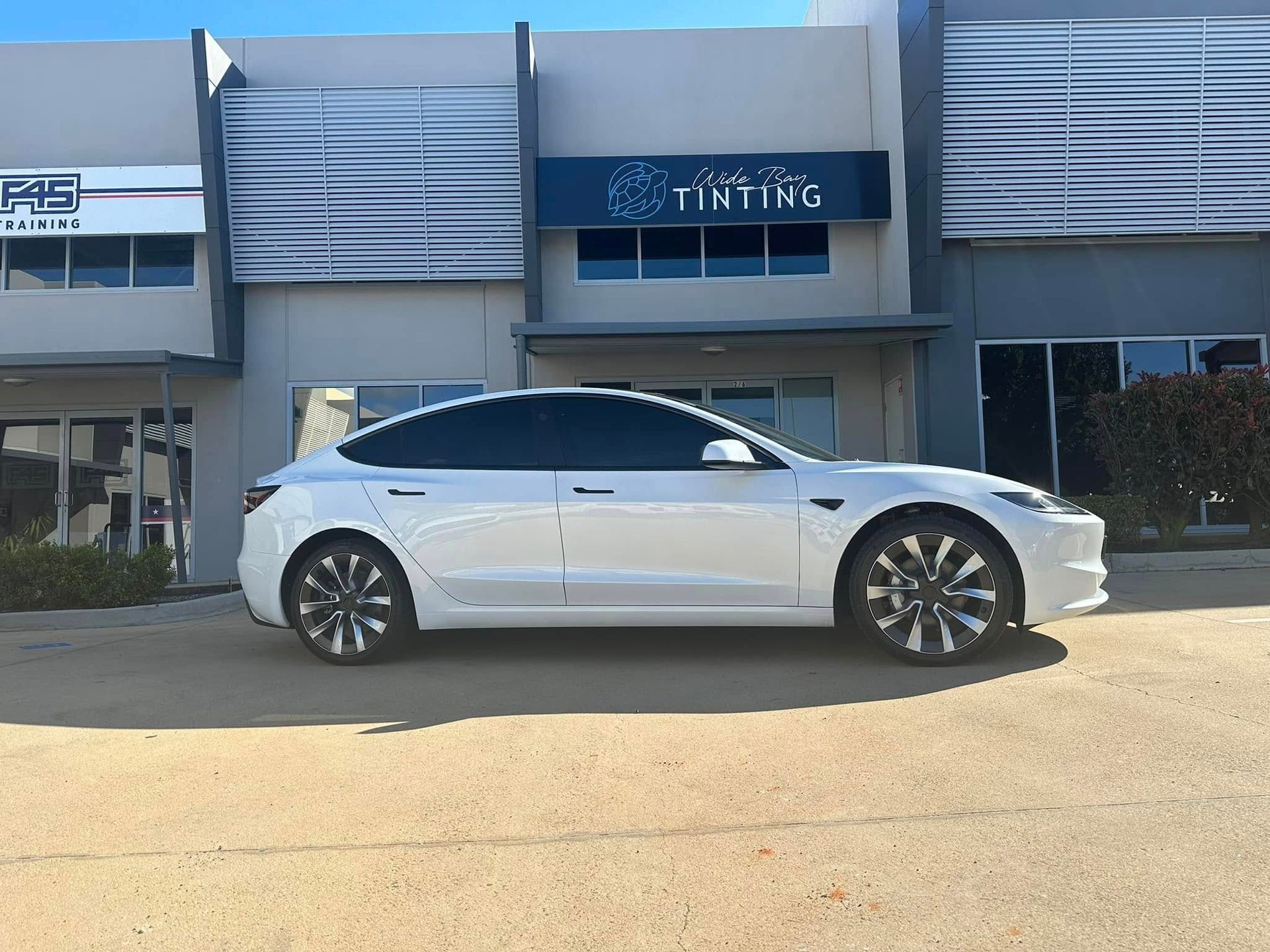 White Tesla Model 3 sedan with tinted windows parked in front of a building with tinting signage — Wide Bay Tinting in Kensington, QLD