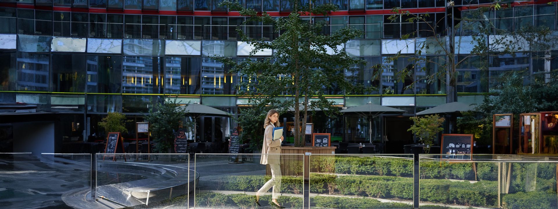 A person walks in front of a building with many windows and greenery.