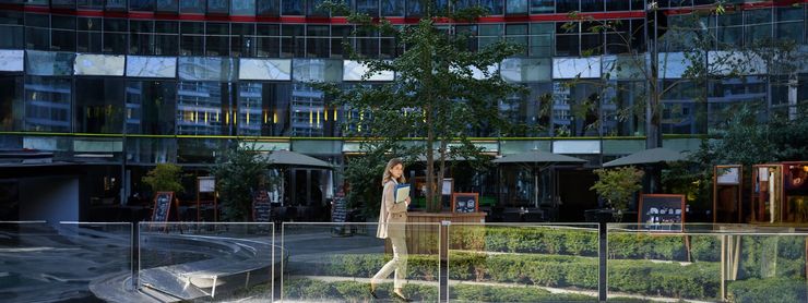 A person walks in front of a building with many windows and greenery.