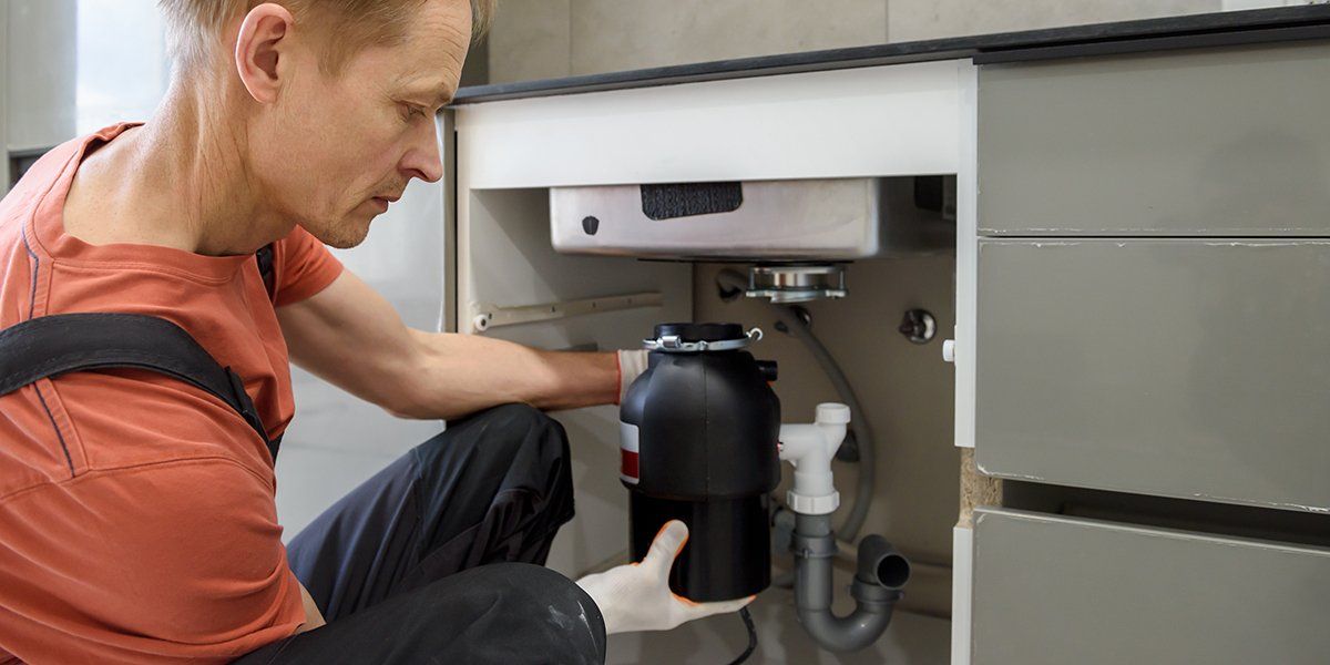 Man installs a garbage disposal under a sink. He is wearing overalls and a red shirt.