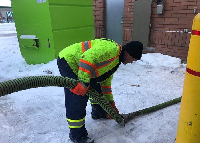 Person in safety gear connecting a large hose on a snowy surface near a grease trap.