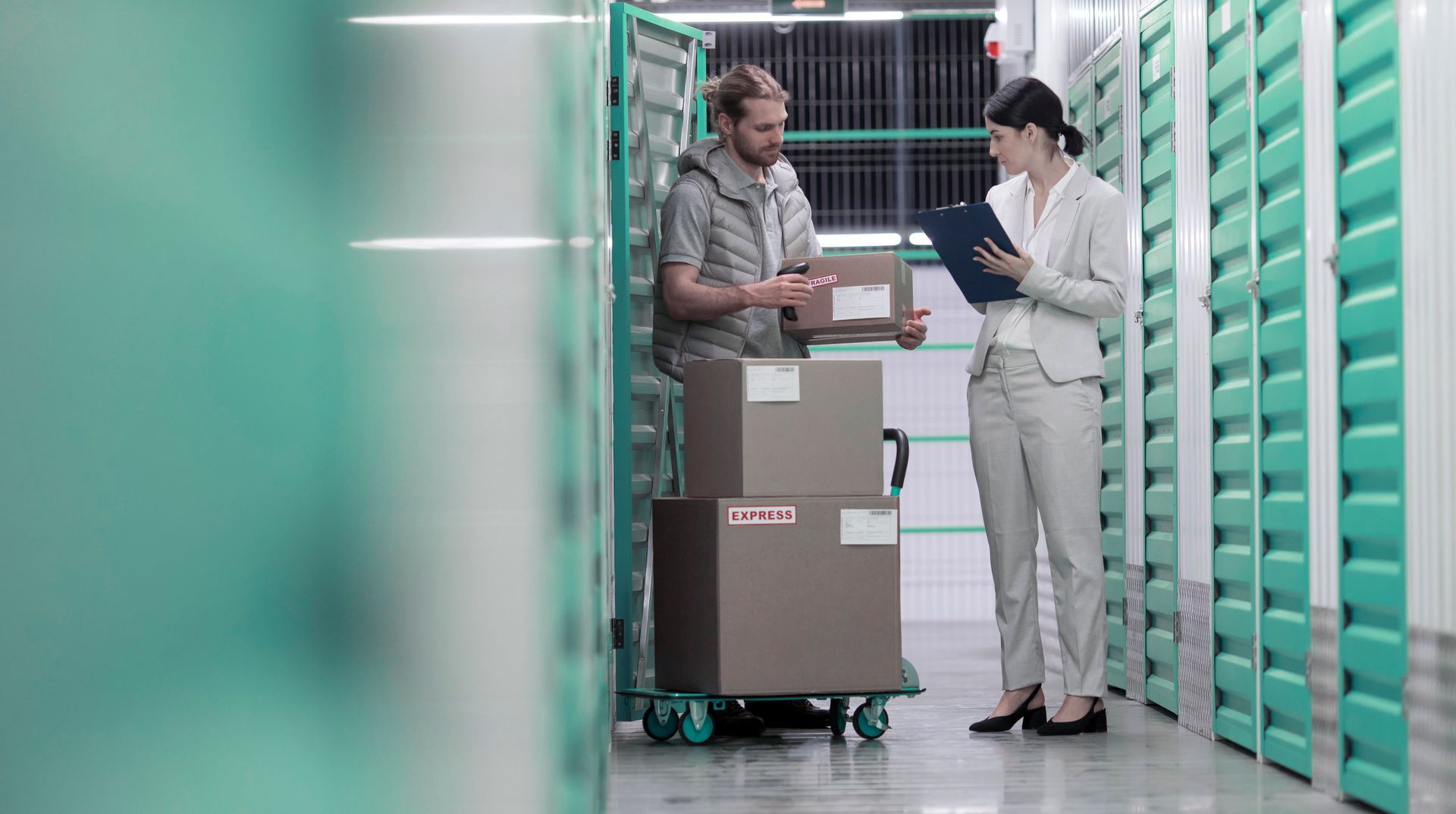We Box Self Storage Um homem empurra um carrinho com caixas enquanto uma mulher olha para uma prancheta.