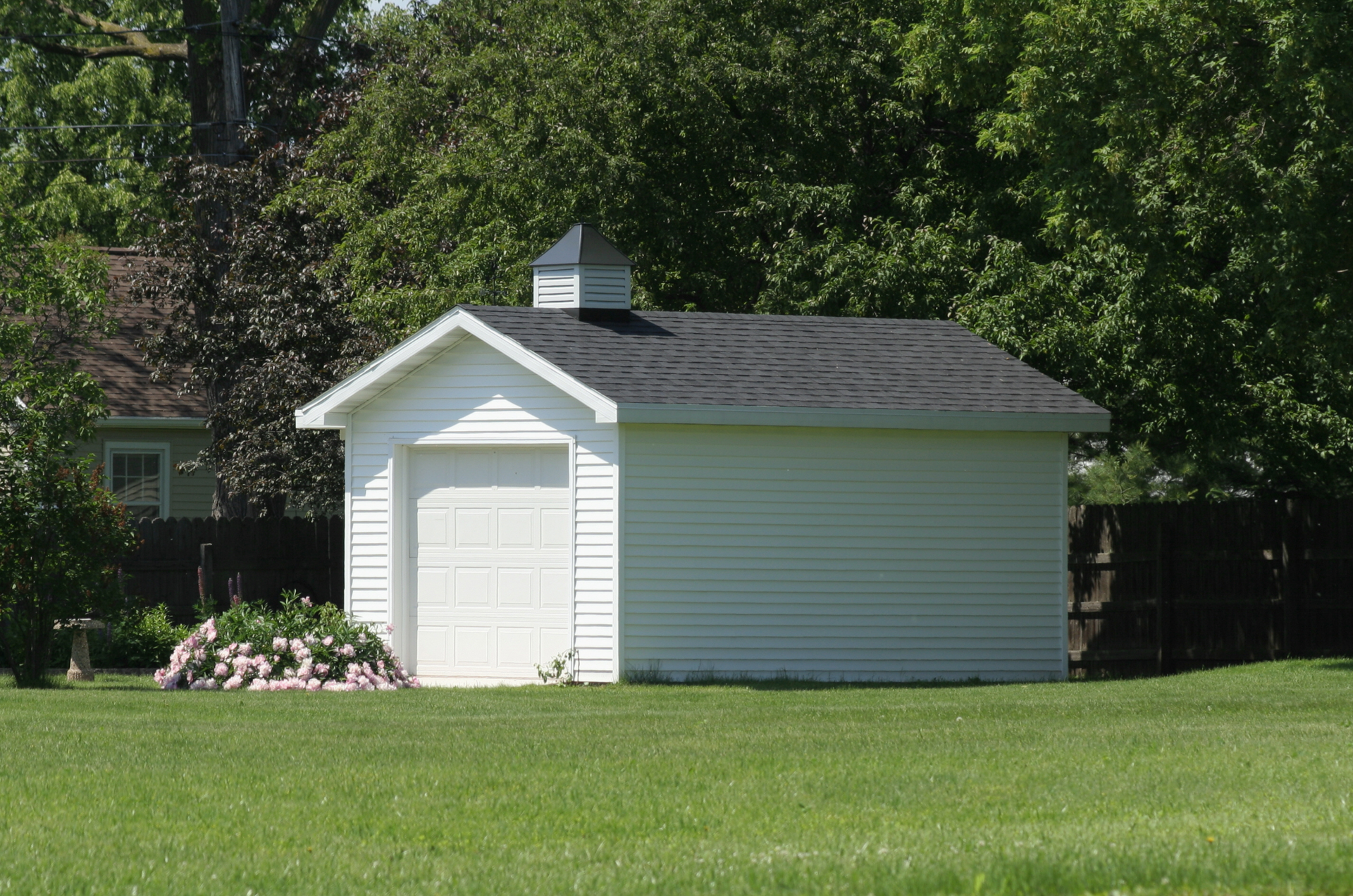 White shed with black roof, small cupola, and garage door on green lawn with trees.